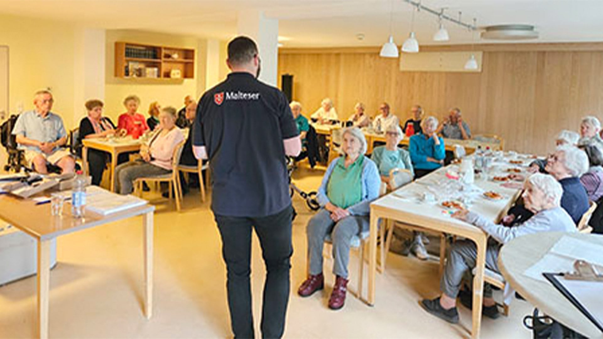 Evdiak Bild von einer Veranstaltung bei der Senioren an unterschiedlichen Tischen sitzen und einem Mann der Malteser zuhören