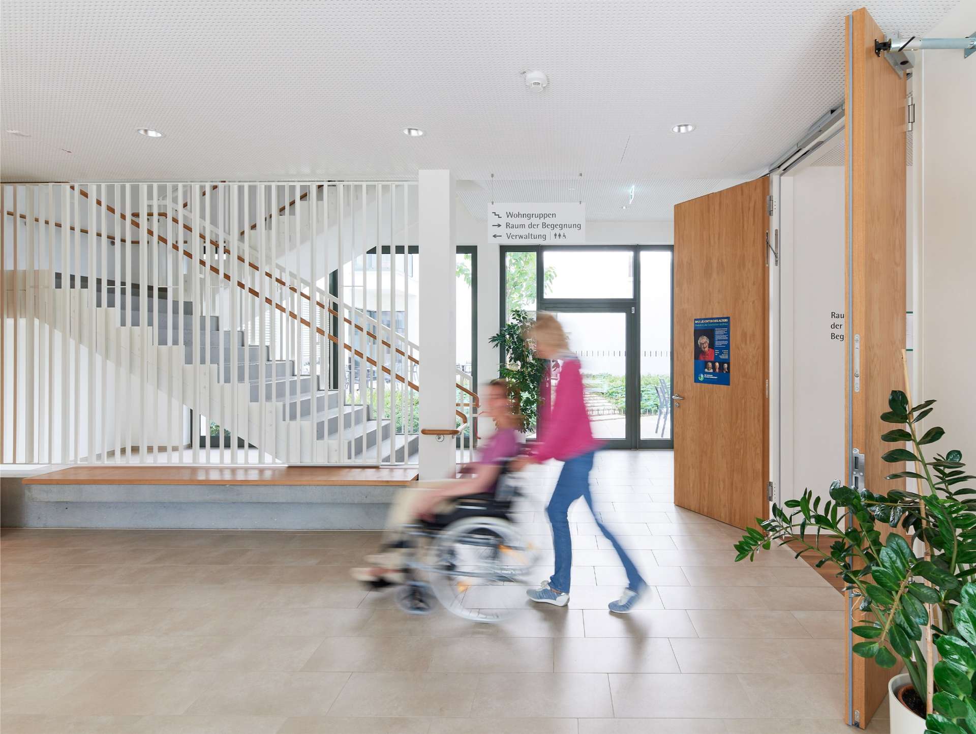 Evdiak Bild von einem Innenraum der Pflegeeinrichtung. Eine Frau schiebt eine andere Frau im Rollstuhl