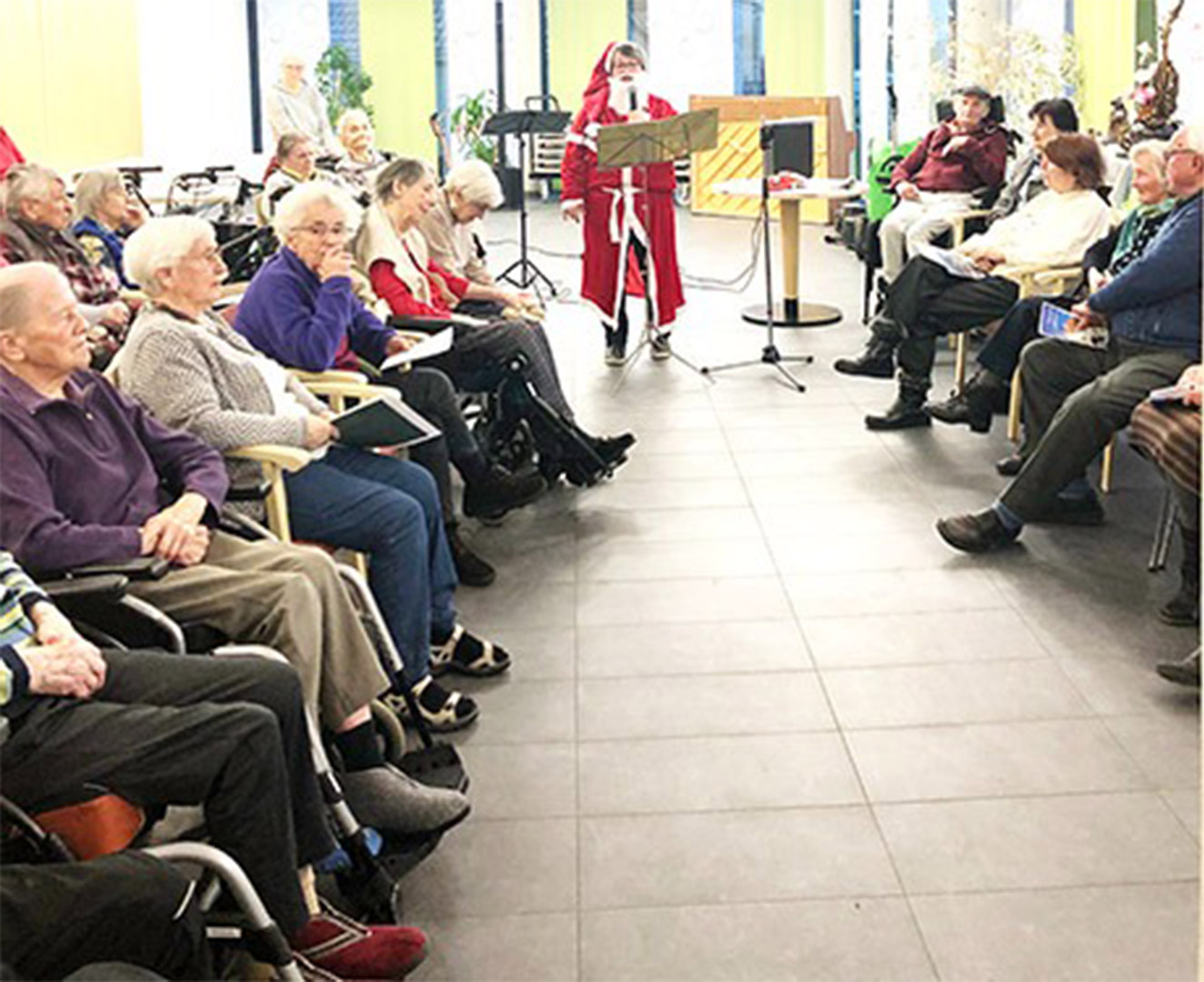 Evdiak Bild einer Frau im Nikolauskostüm, die ein Mikrophon spricht. Senioren schauen ihr dabei zu.