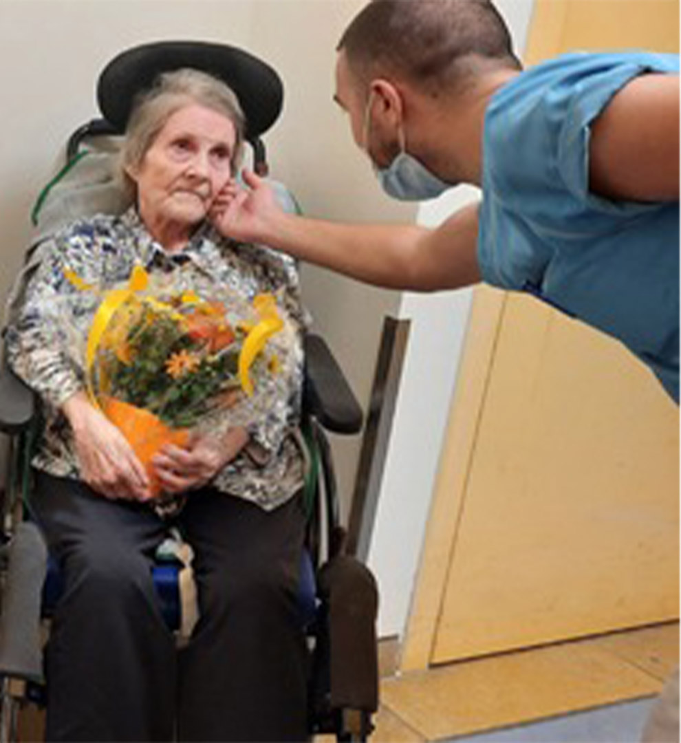 Evdiak Bild von einer Seniorin im Rollstuhl. Ein Pfleger streicht ihr über die Wange und sie hält einen Blumenstrauß