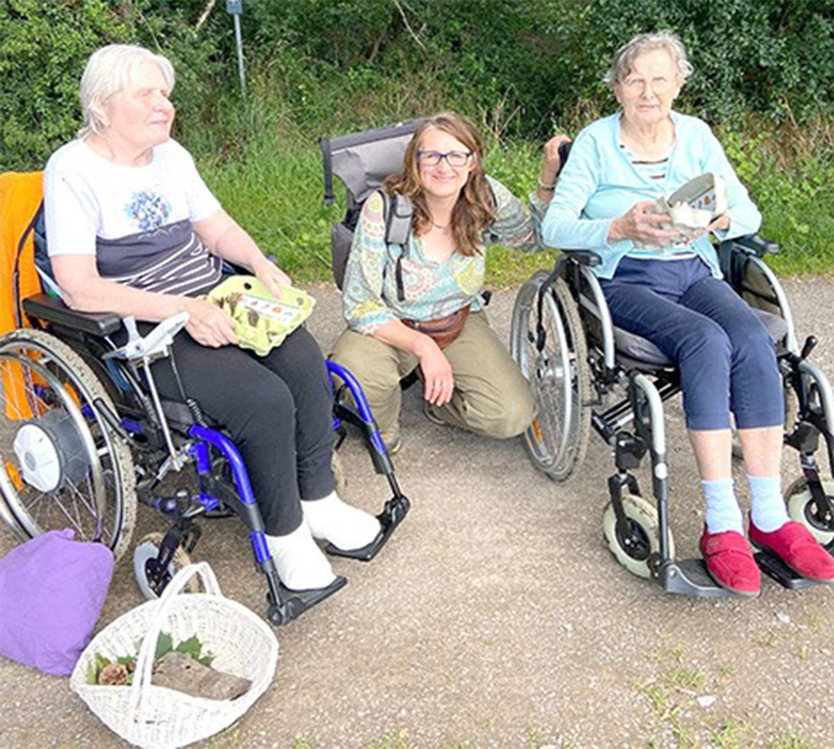 Evdiak Bild von zwei Seniorinnen in Rollstühlen und einer Mitarbeiterin zwsichen ihnen