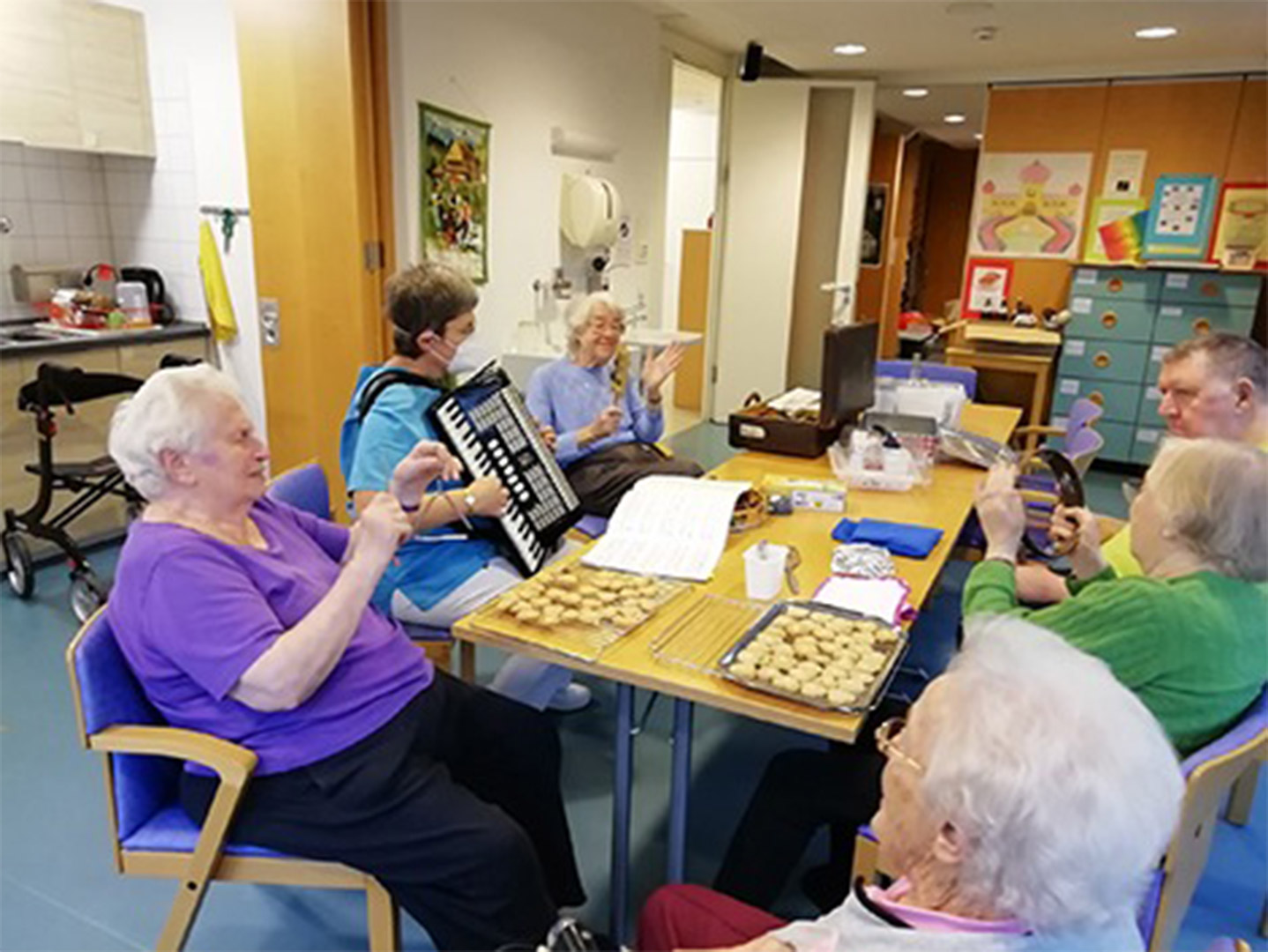Evdiak Bild von einem Tisch mit Senioren. Auf diesem liegen Plätzchen und es wird musiziert