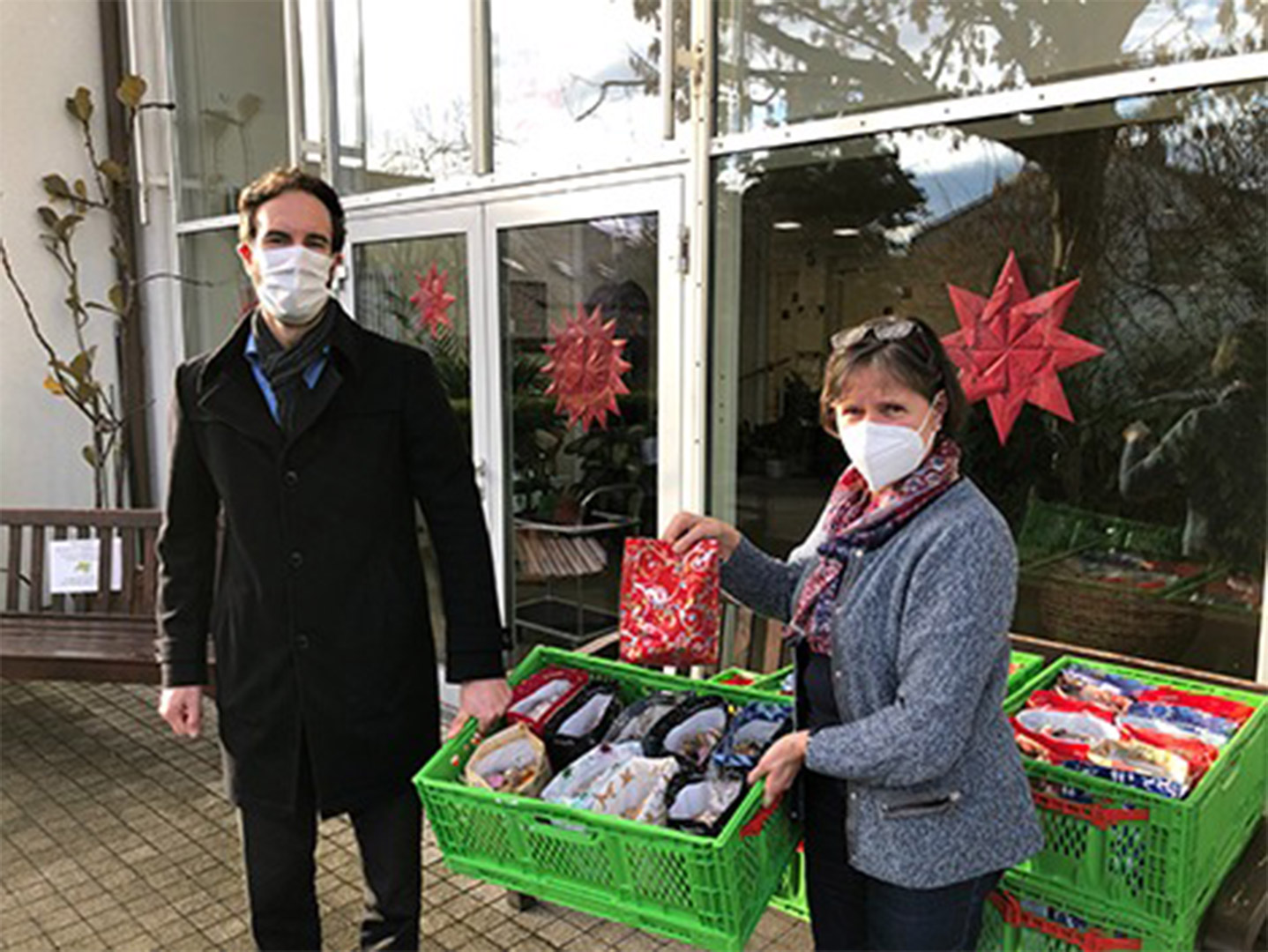 Evdiak Bild von zwei Personen mit Coronamasken. Sie tragen eine Kiste mit vorgepackten Tüten zwischen sich.