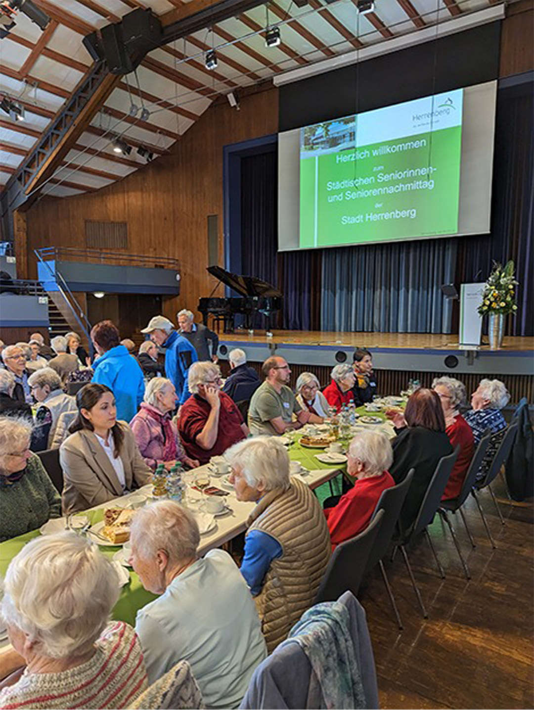 Evdiak Bild von Senioren, die an langen Tischen sitzen, im Hintergund ist eine Bühne