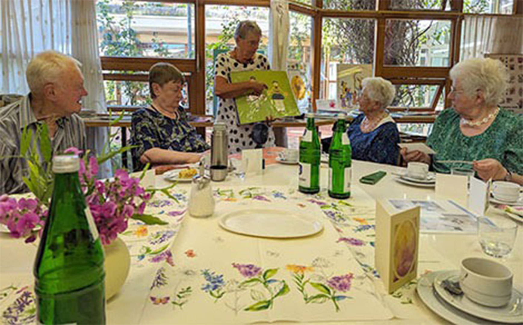 Evdiak Bild von 5 Senioren an einem Tisch, eine Seniorin steht und zeigt ein Kunstwerk