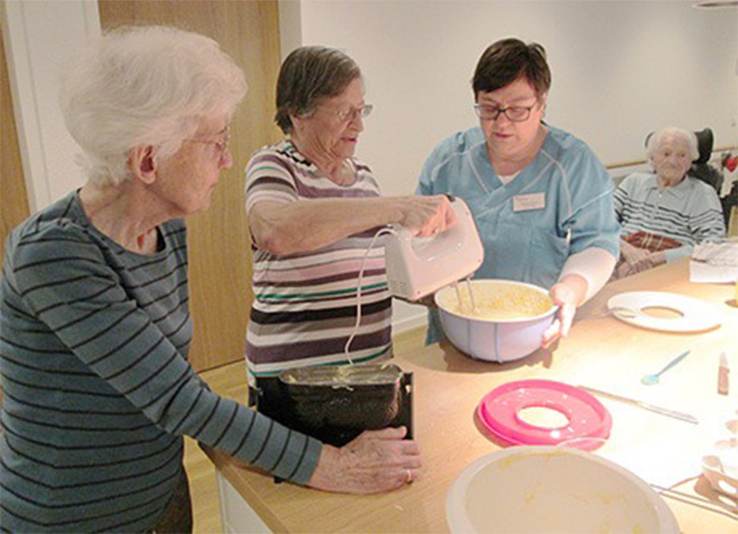 Evdiak Bild von zwei Seniorinnen und einer Mitarbeiterin. Eine Seniorin bedient mit der Mitarbeiterin ein Handrührgerät