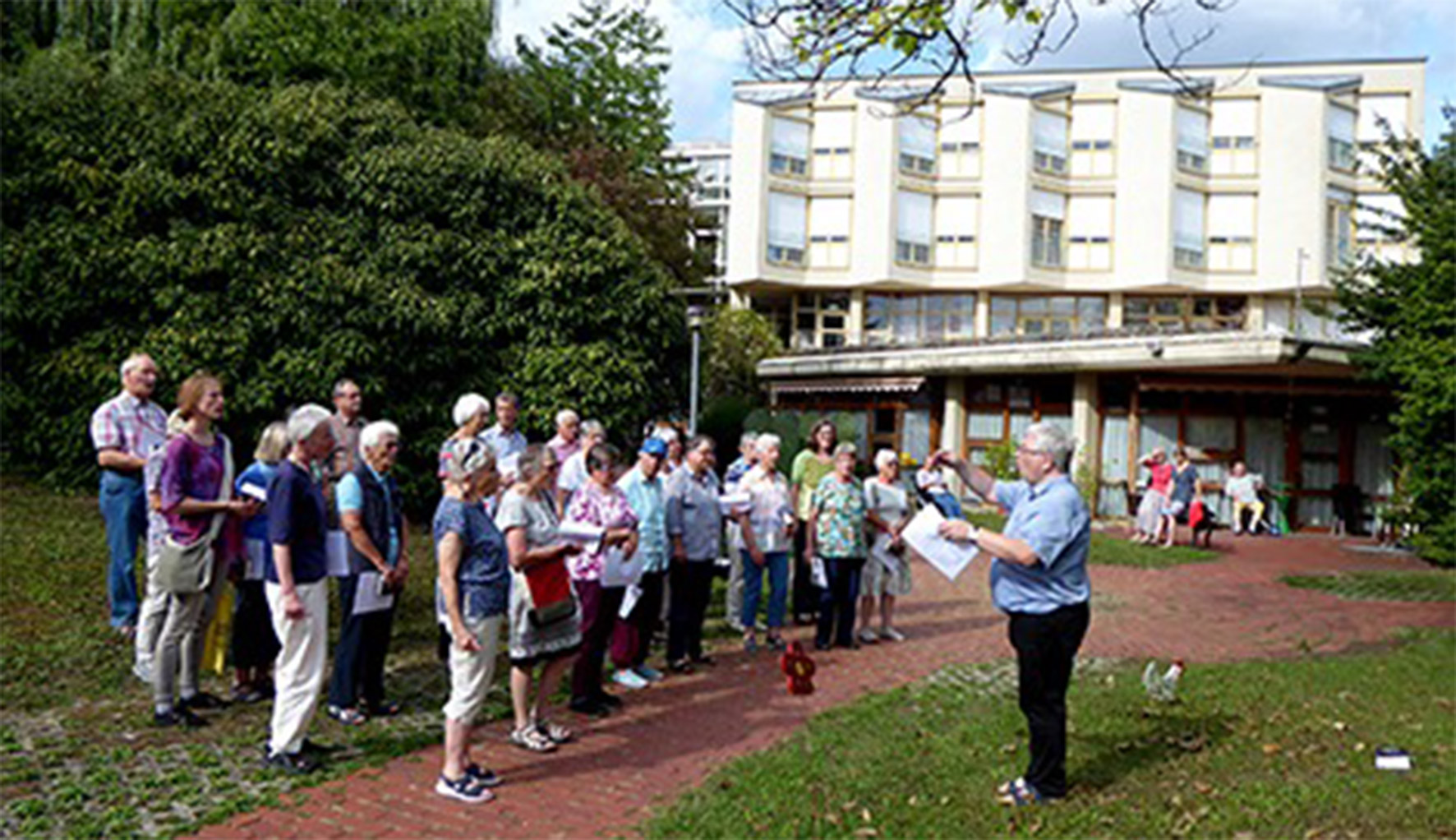 Evdiak Bild von einem Seniorenchor und ihrem Dirigent, die im Freien musizieren.