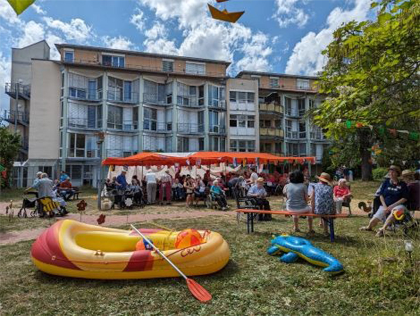 Evdiak Bild von einem Sommerfest. Im Vordergrund liegt ein Schlauchboot, im Hintergund sitzen Senioren unter einem Pavilion.