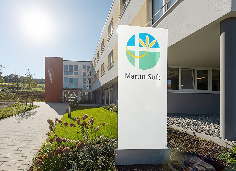 Evdiak Im Vordergrund, Schild mit Logo der Einrichtung und im Hintergrund das Gebäude